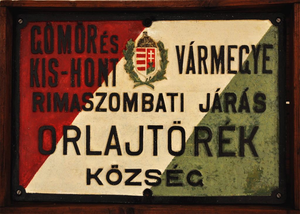 Tabuľa označujúca názov obce Malé Teriakovce z obdobia Rakúsko-Uhorska. V drevenom ráme je trojfarebný červeno-bielo-zelený podklad, uprostred  je opatrená erbom Uhorska.  Jej rozmery sú 53 x 38 cm.